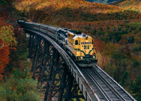 A freight train crosses a wooden trellis bridge over a western landscape.